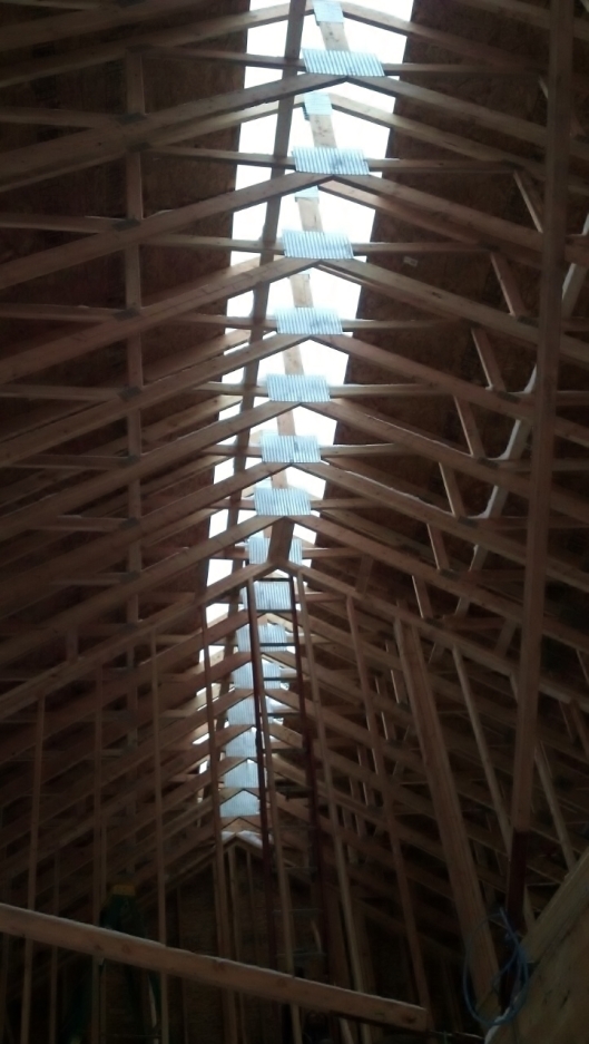 view of roof framing of a house under construction. The roof is sheated almost up to the top leaving open a section on each of the peak where light comes through.