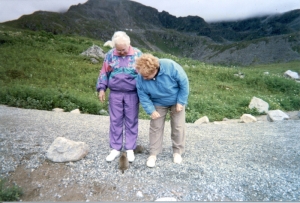 two mature women standing on a path in front of rugged mountains looking at a marmot sitting at their feet.