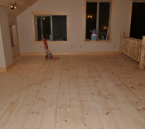 looking across room at newly sanded, unfinished pine floor boards with sanding machine at the far left end of the room.